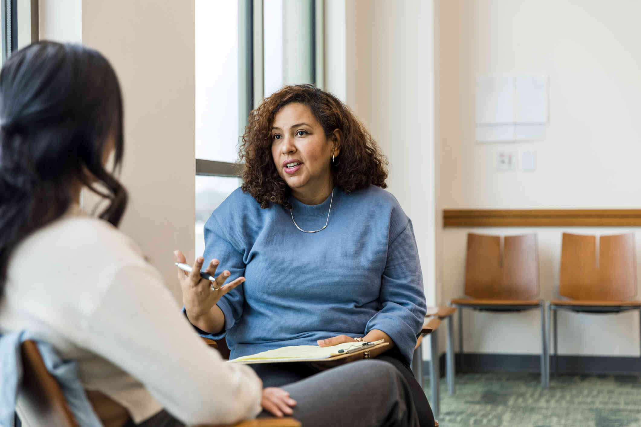 A female therapist in a blue sweater sits across from her female patient and talks to her during a therapy session.